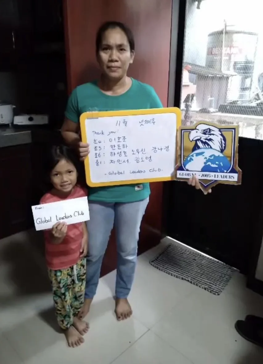 A woman and a young girl standing indoors, holding signs related to the Global Leaders Club. The woman is holding a whiteboard with handwritten notes and a digital badge, while the girl holds a paper with the club's name. The background shows a windo