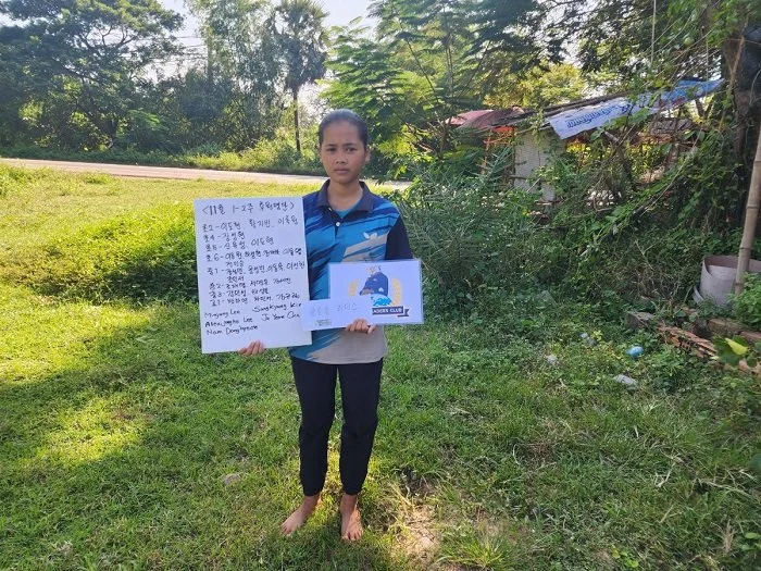 A young girl holding informational signs on a grassy area with trees and makeshift structures in the background.