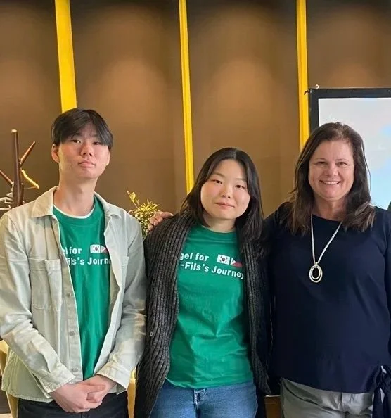 Three people standing indoors, two women and one man, all smiling. The man and woman in the middle are wearing green t-shirts with white text. The woman on the right is wearing a black top and necklace. Yellow vertical beams are in the background.