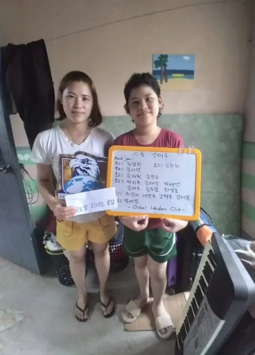 Two children standing indoors, holding signs and smiling. The child on the left holds a card with an eagle graphic, and the child on the right holds a whiteboard with handwritten text. There is a painting of a beach scene on the wall behind them, wit