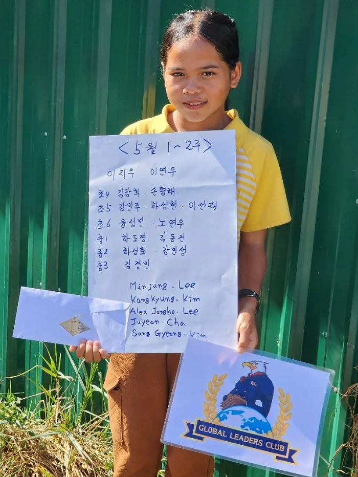 A young girl in a yellow shirt holding a handwritten list of names in Korean and English, standing in front of a green metal fence. She is also holding a card with the Global Leaders Club logo, featuring an eagle perched on a globe surrounded by a la