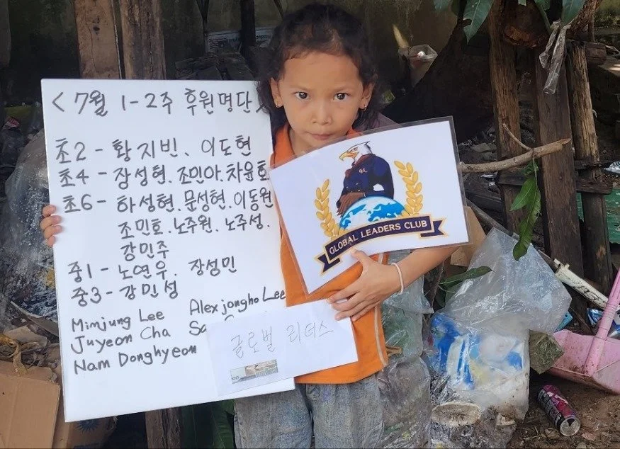 Young girl standing outdoors holding a sign with a list of names and a logo of the Global Leaders Club. The background shows clutter and debris.