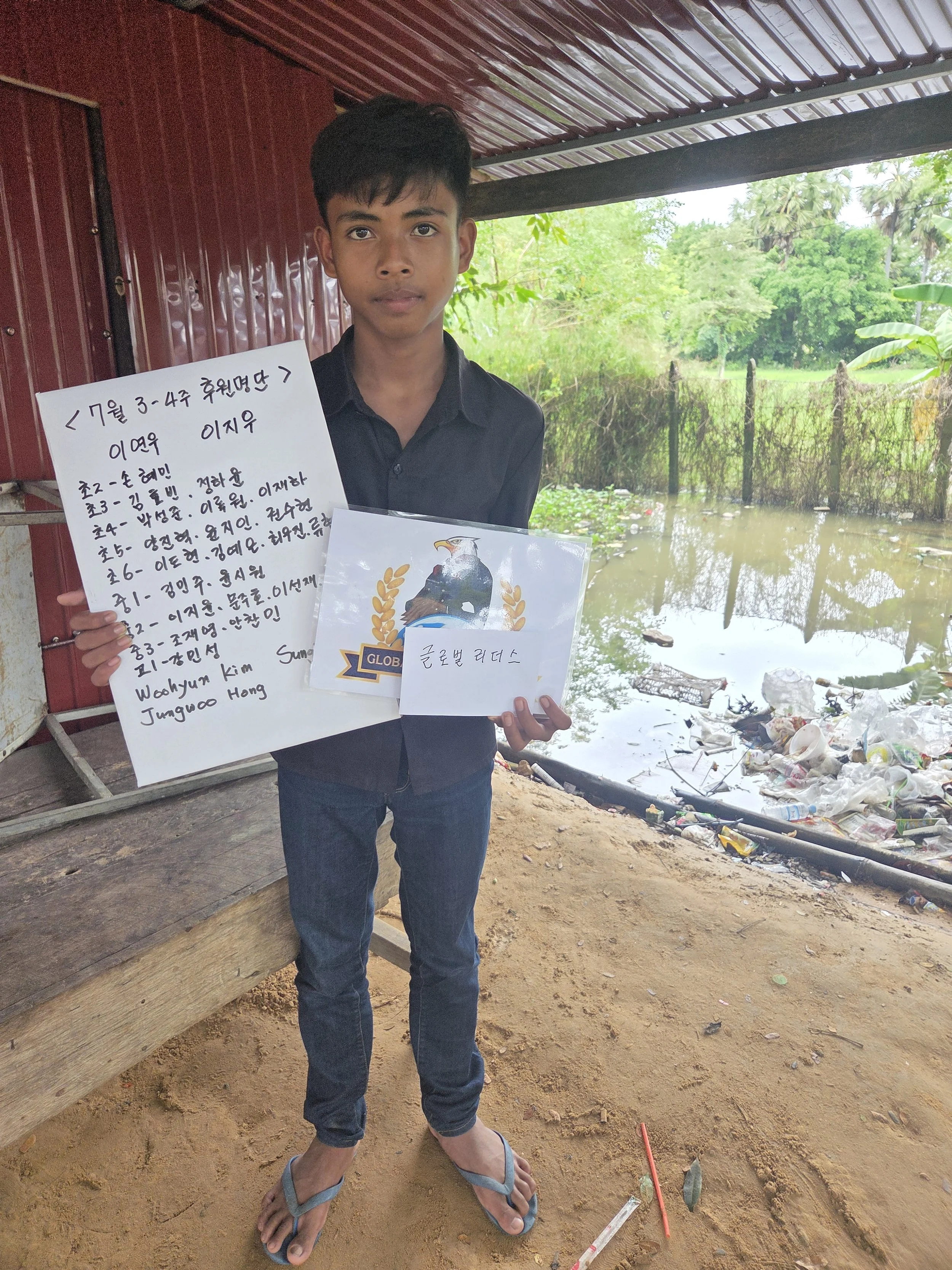 A young boy holding handwritten notes and a poster in front of a flooded area with trash and algae, under a roof with green trees in the background.