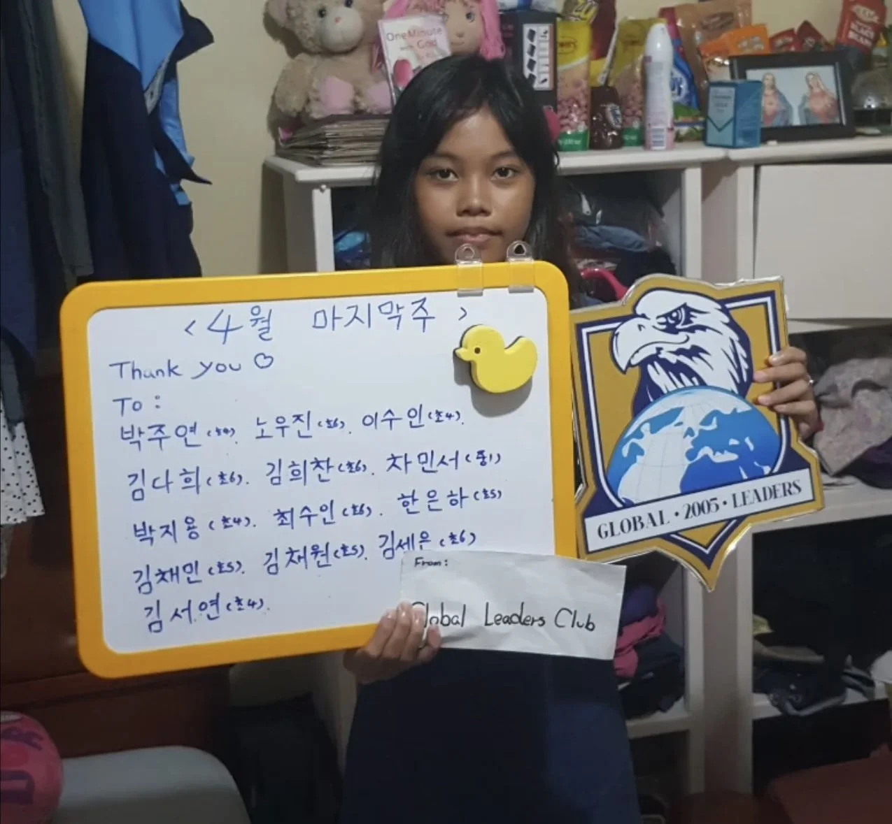 A young girl with long dark hair holding a whiteboard and a badge. The whiteboard has handwritten Korean and English text thanking people, decorated with a yellow duck magnet. The badge features an eagle's head above a globe with the words "GLOBAL 20