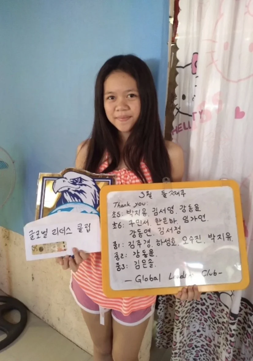 A young girl standing indoors, holding a handwritten sign with Korean writing, a smaller card with some Korean writing and a sticker of an eagle or hawk, and a decorated box with Korean writing. She is wearing a red and white striped top and pink sho