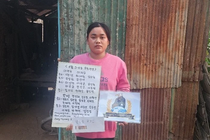 A young woman holding a handwritten sign and a printed document in a rural setting with a corrugated metal wall in the background.