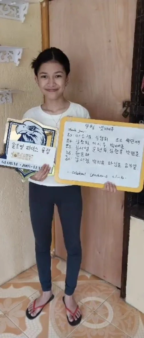 A young girl is standing indoors, holding a sign with Korean writing and a logo with an eagle's head. She is smiling, wearing a white shirt, dark pants, and flip-flops, with a yellow frame around the sign she is holding.