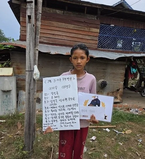 A young girl with dark hair, wearing a pink shirt and red pants, standing outdoors in front of a wooden and corrugated metal house, holding a handwritten sign and an illustrated paper.