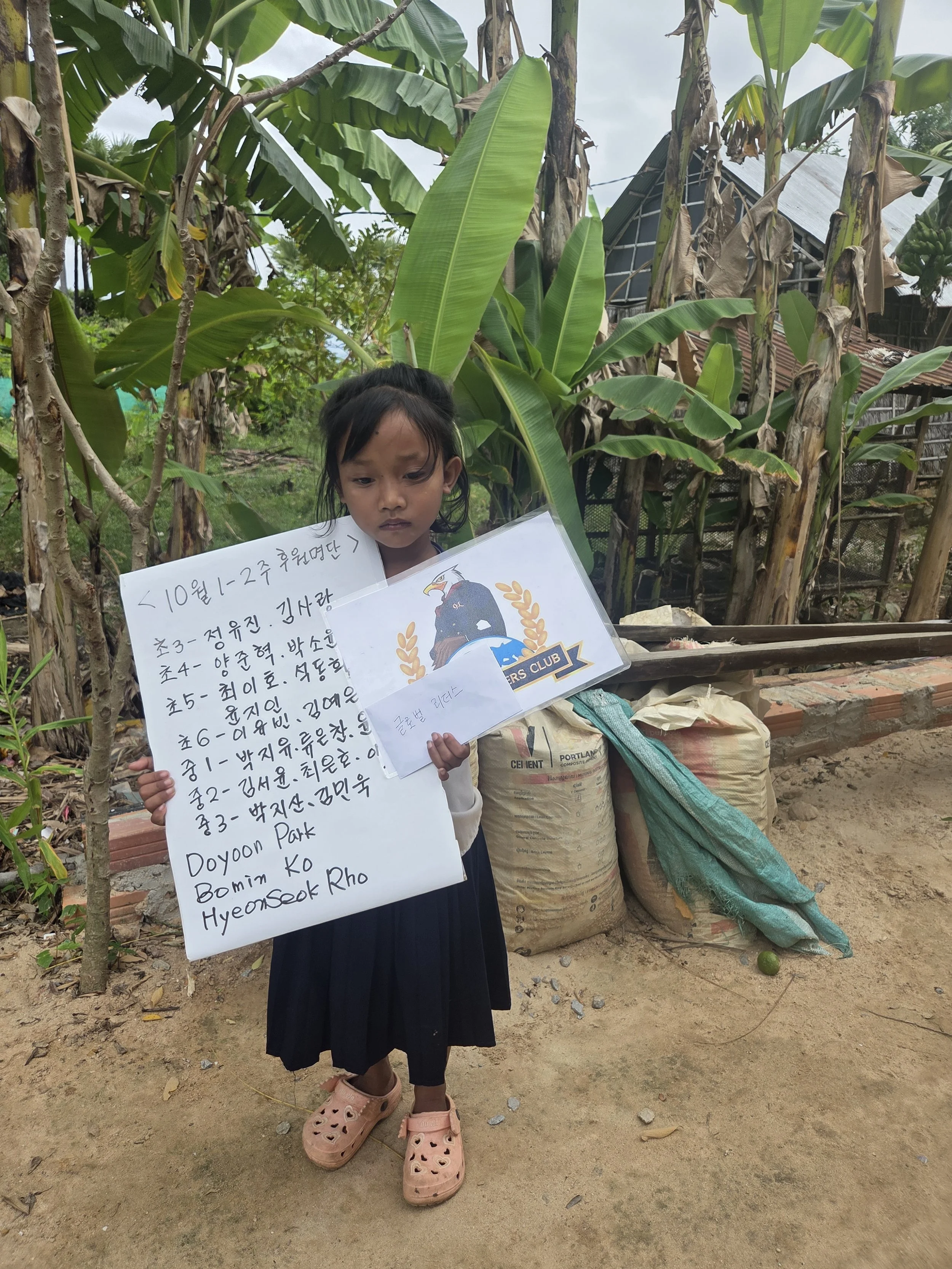 A young girl standing outdoors in front of banana plants, holding two sheets of paper with handwritten messages and a printed emblem. She is wearing a dark dress, pink Crocs, and has dark hair.