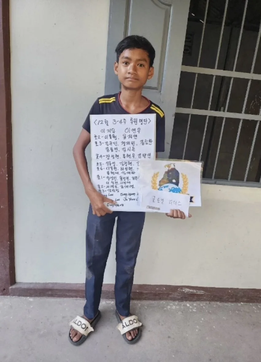 A young boy standing indoors near a barred window, holding a handwritten poster with text and a drawing of a military emblem.