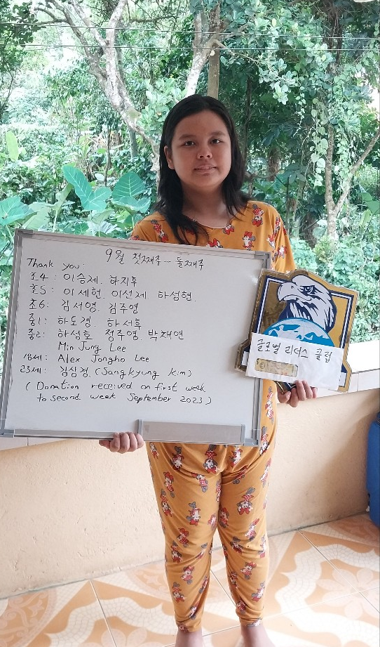 A girl wearing yellow pajamas with a cartoon character pattern, standing outside on a tiled floor, holding a whiteboard with writing in Korean and English, and a sign with an eagle logo, in front of a background of green trees and plants.