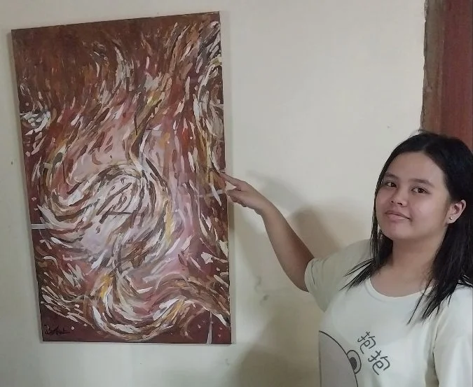 A young woman with long black hair in a white shirt stands indoors pointing at an abstract painting on a wall.