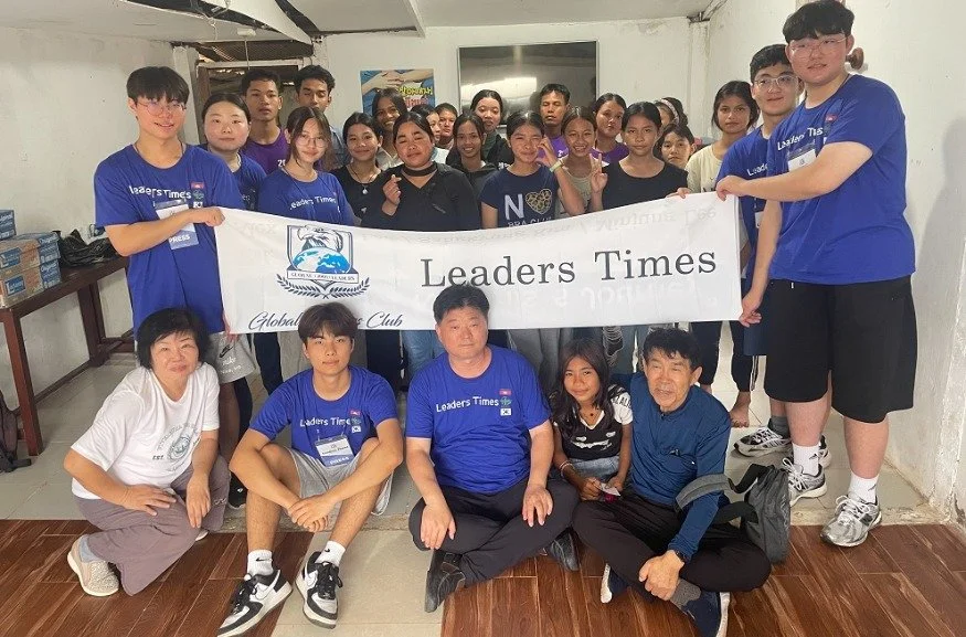 A group of young people and adults holding a banner that reads 'Leaders Times' at an indoor event, with some individuals wearing matching blue t-shirts with the same logo.