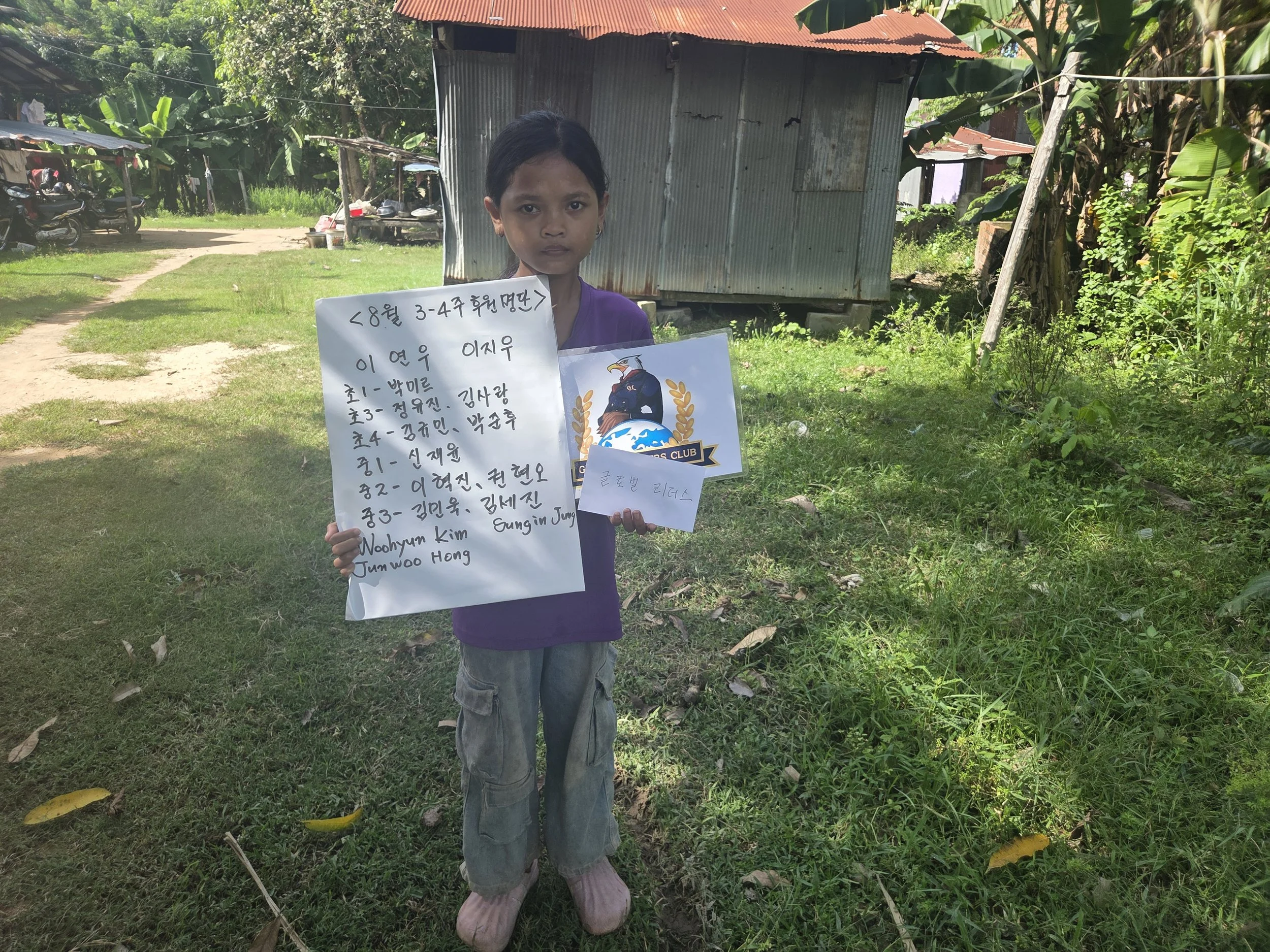 A young girl standing outdoors, holding a whiteboard with handwritten text in Korean and English, and a sign with a logo. She is wearing a purple shirt and gray pants. Behind her is a grassy area with a small, rustic metal building, trees, and motorc
