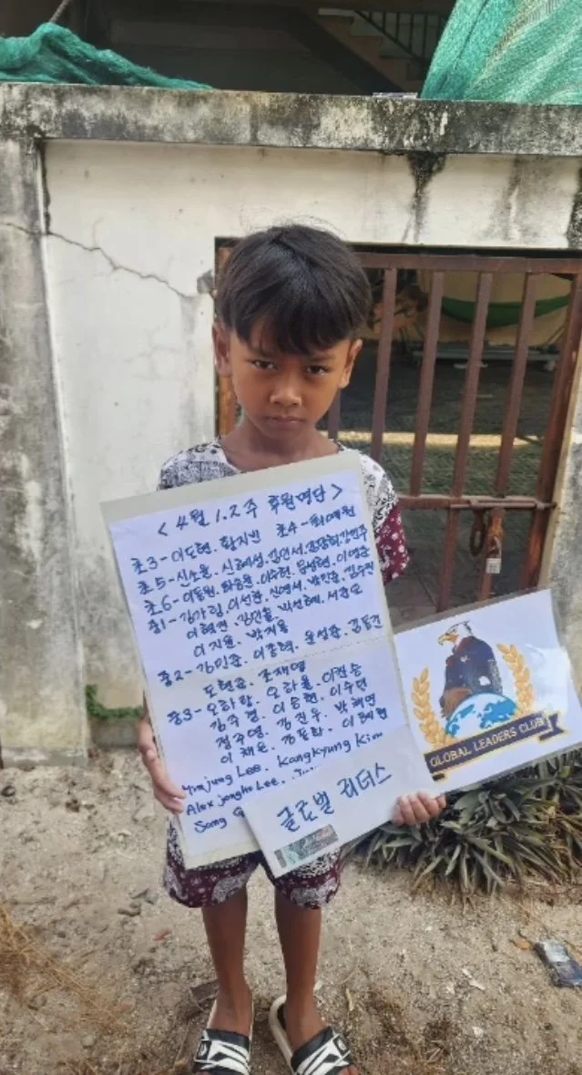 A young boy with short dark hair holding a handwritten sign in a foreign language, standing outside in front of a concrete wall and metal gate, with a sticker that says 'Global Leaders Club' and a small flag underneath.