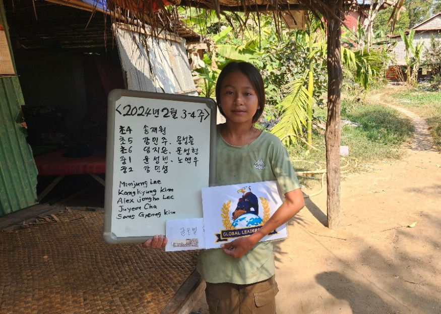 A young girl standing outdoors holding a whiteboard with handwriting and a booklet. The background shows a rural setting with banana trees and a dirt path.