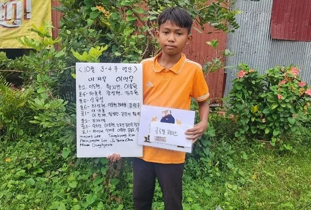 A young boy with short black hair wearing an orange polo shirt and dark pants standing outdoors, holding a white sign with handwritten text and a book cover with a dog illustration, in front of greenery and a weathered fence.