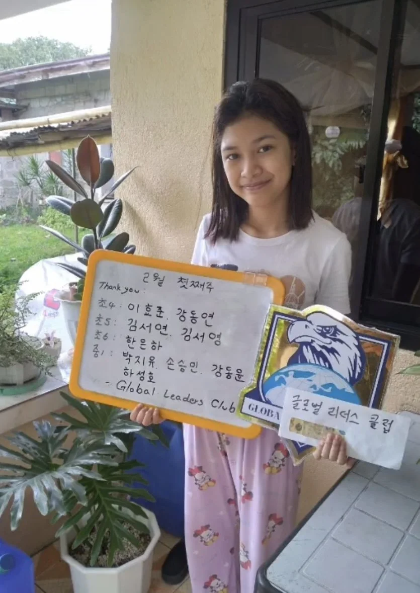 A young girl with shoulder-length dark hair, wearing a white shirt and pink pajama pants with character prints, stands outside near a window, holding a whiteboard and a sign with a blue and white logo depicting an eagle and a globe. The whiteboard ha