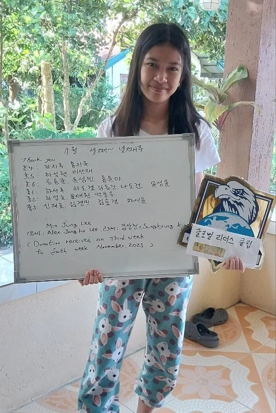 A girl standing outdoors on a porch holding a whiteboard with writing on it and a certificate with an eagle logo. She is wearing pajama pants with bunny prints and a white t-shirt, standing in front of a pink wall, with shoes on the floor nearby and 