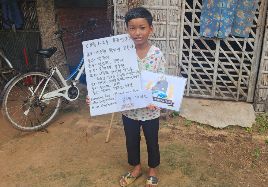 A young boy standing outdoors on dirt ground, holding a whiteboard with writing and a flag, in front of a bamboo fence and a wooden structure. A bicycle is leaning against the fence.
