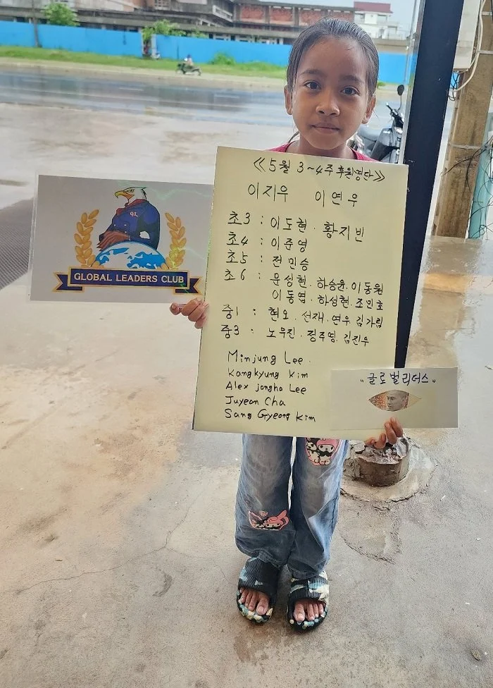 A young girl standing outside holding a sign with handwritten Korean names and some English names. There is a sign for 'Global Leaders Club' next to her. The background shows a wet road and some buildings.
