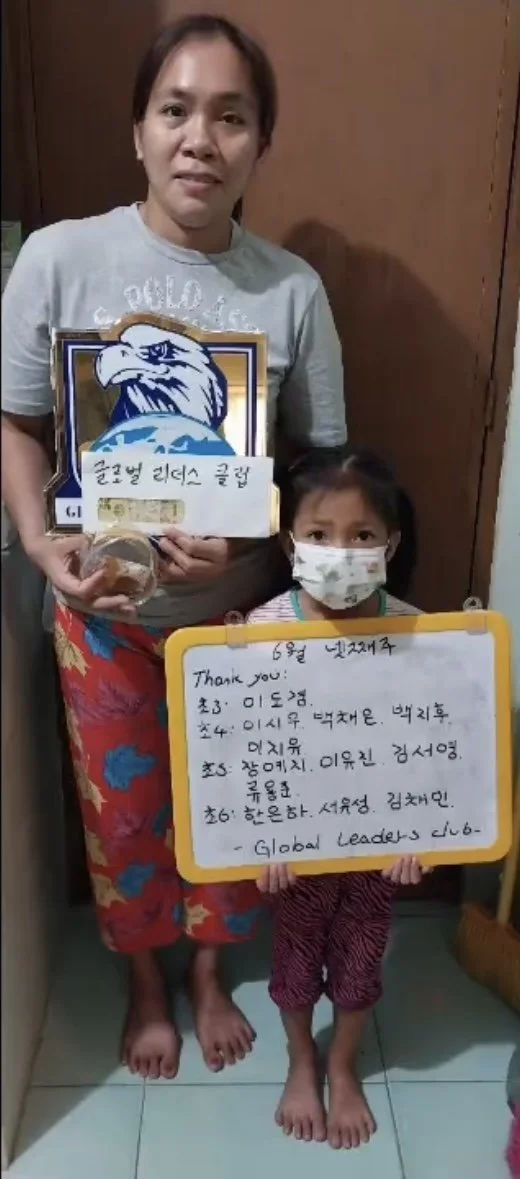 A woman and a girl standing indoors. The woman is holding a plaque with an eagle logo and Korean text. The girl is holding a whiteboard with handwritten notes and is wearing a face mask. Both are barefoot.