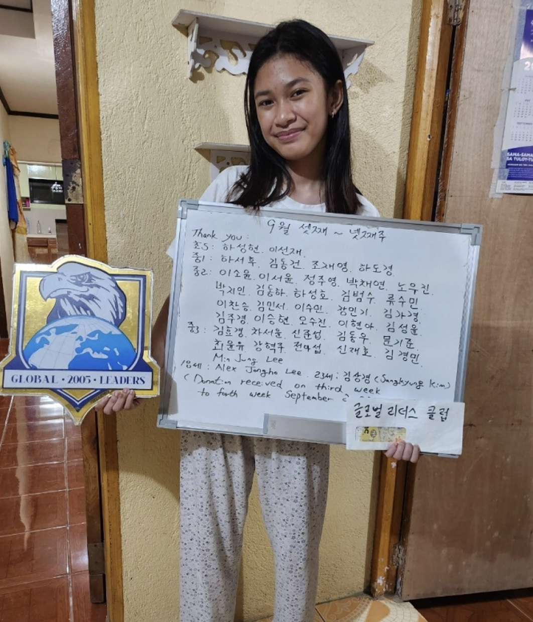 A young girl with dark hair holding a plaque and a whiteboard with handwritten text, standing inside a room with yellow walls and a wooden door.