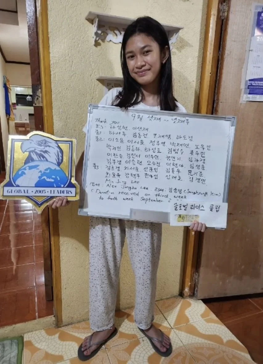 A young woman standing indoors, holding a whiteboard with handwritten text in Korean and English, and a shield-shaped sign with an eagle logo that reads 'Global 2005 Leaders.' She is wearing pajamas and flip-flops.