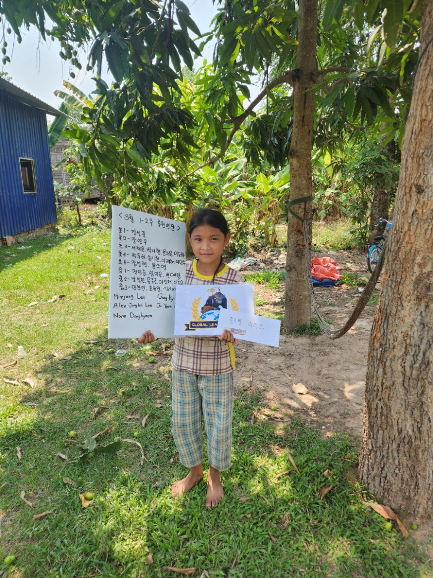 A young girl standing outdoors on grass, holding a sign with a picture of an eagle and a handwritten note in Korean. She is barefoot, wearing a plaid shirt and plaid pants, with trees in the background and a small blue building nearby.
