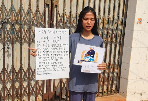 Young woman standing outdoors holding a handwritten sign and a poster, with a metal gate behind her.