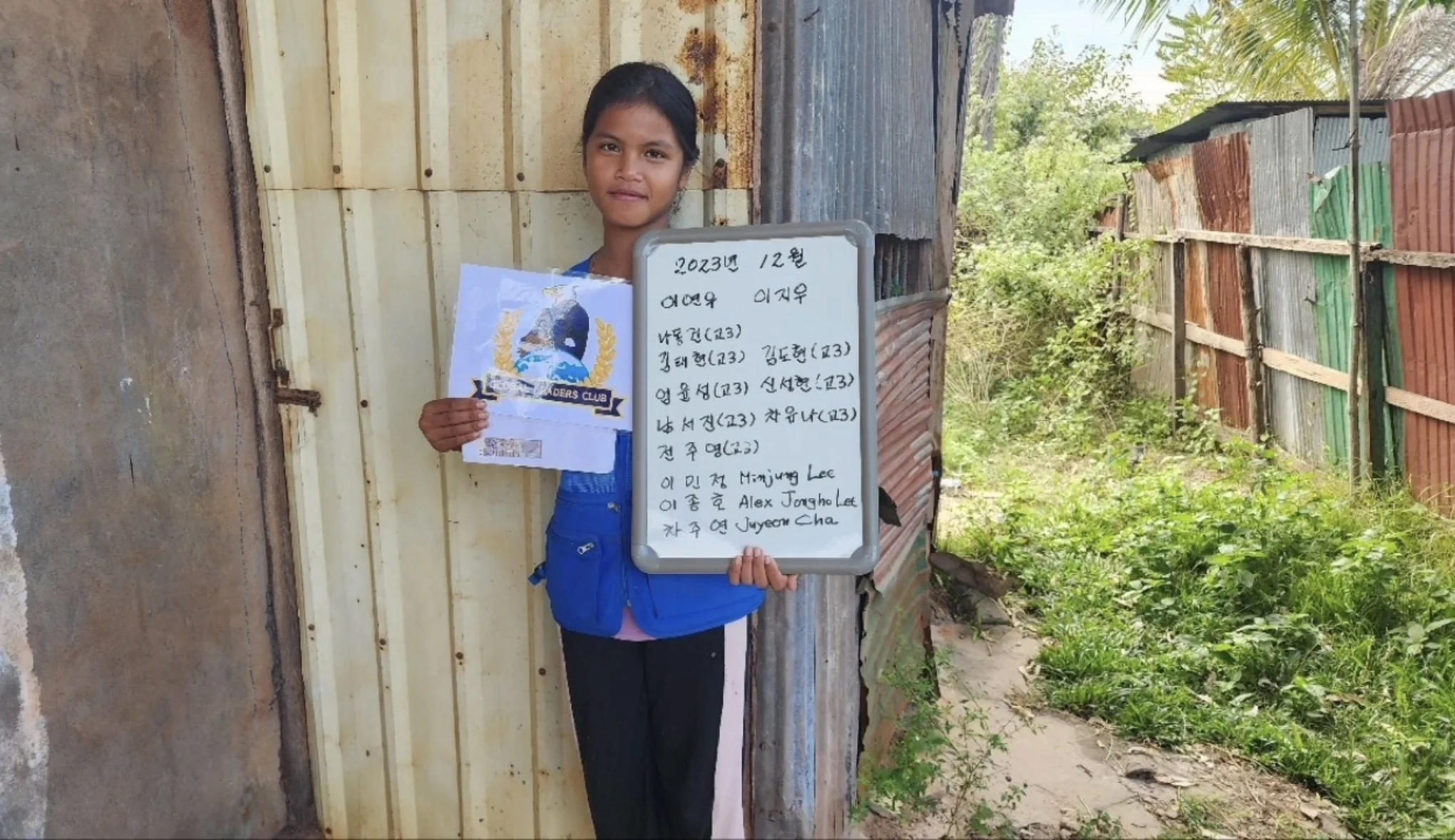 A young girl standing outdoors near a rustic fence and holding a whiteboard with writing in Korean, while also holding a piece of paper with a logo and text. She is wearing a blue jacket and black pants.