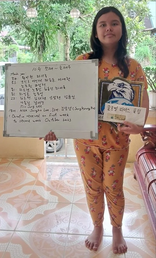 A young girl with long dark hair standing on a tiled floor holding a whiteboard with Korean and English writing, and a paper with an eagle illustration. She is wearing orange pajamas with a pink and white floral pattern, and is barefoot. There is a w