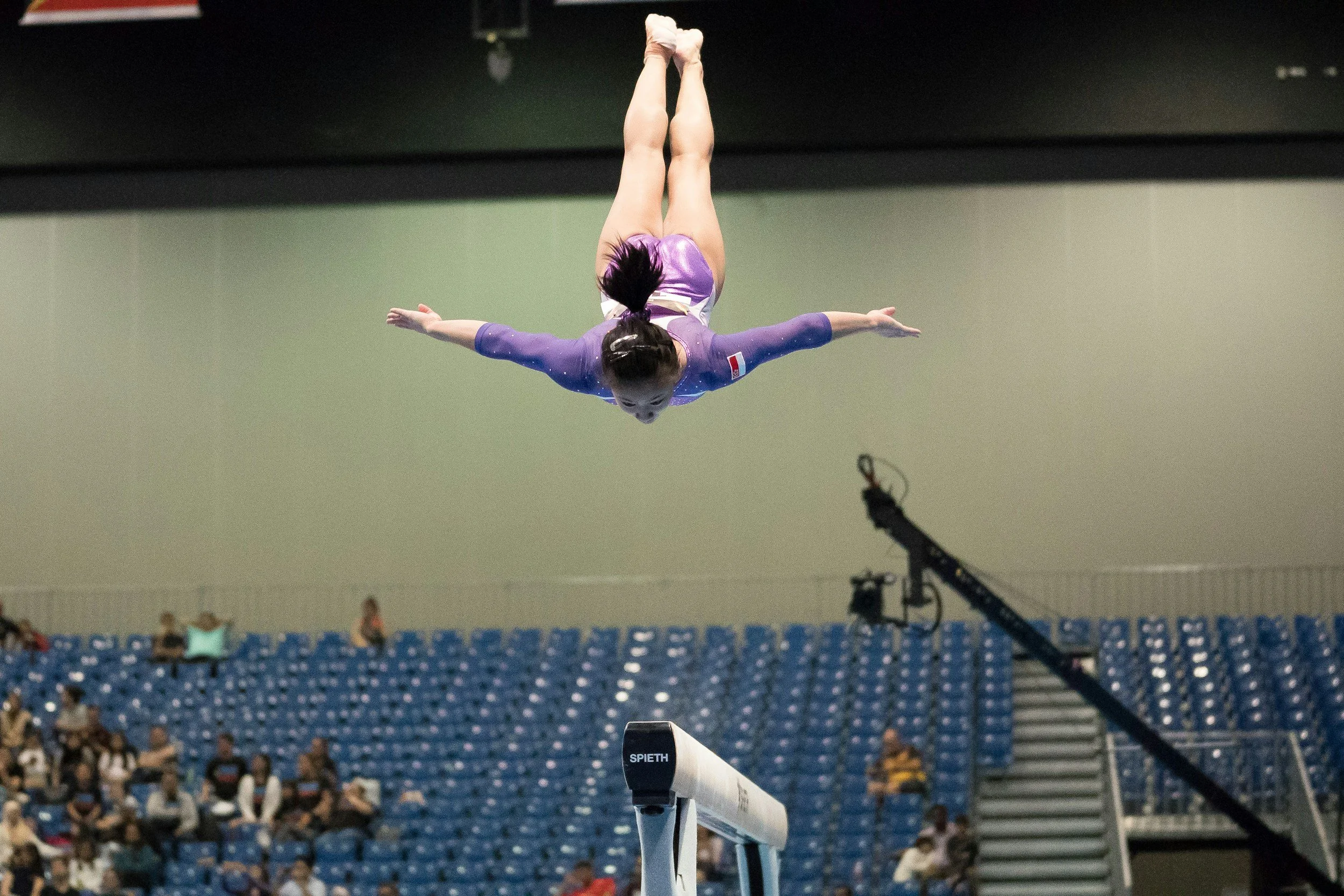 A female gymnast performing a balance beam routine in an indoor sports arena with an audience in the background.