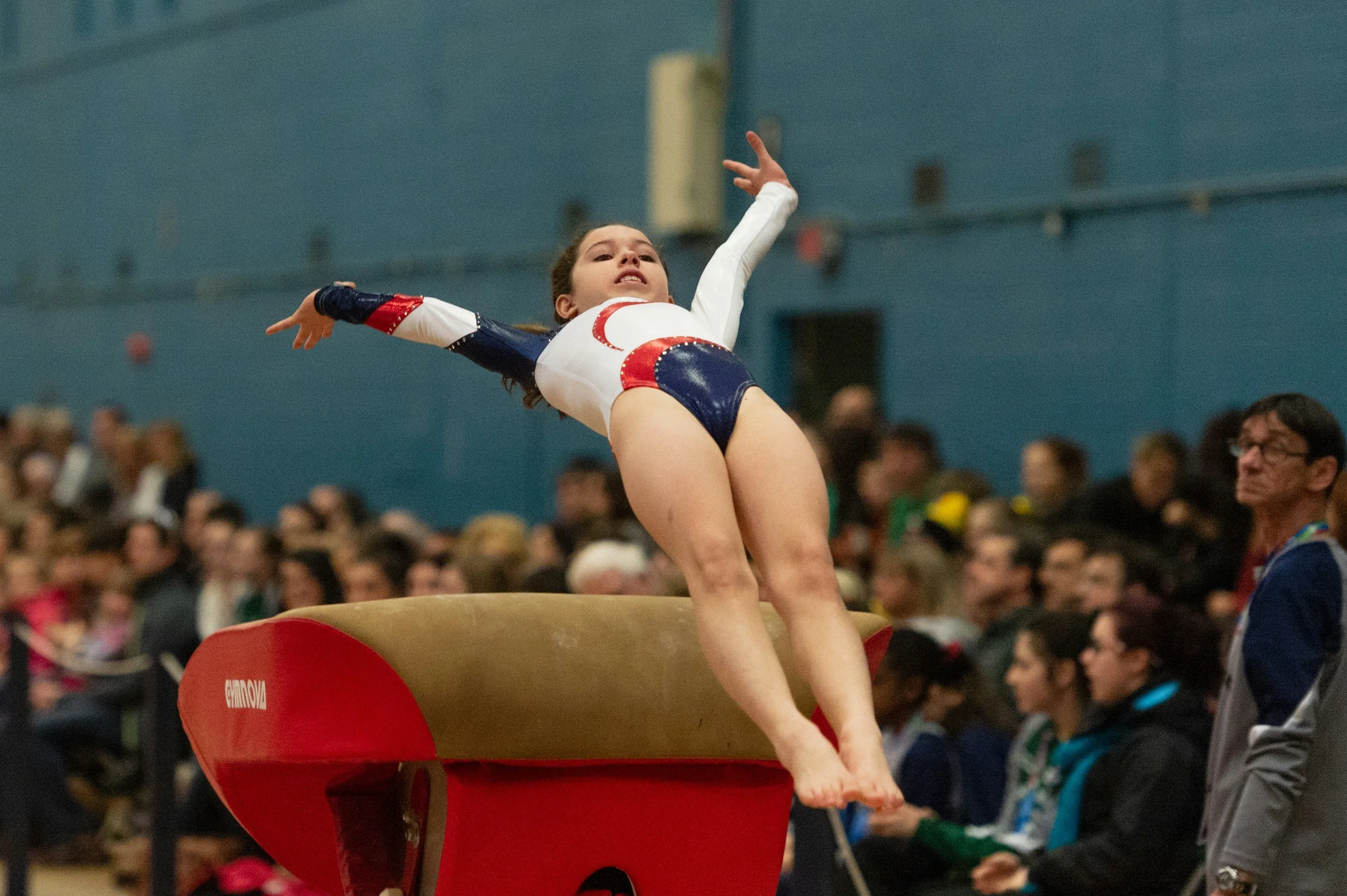 Young female gymnast performing on vault, mid-air with arms outstretched and focused expression, in a gymnasium with spectators in the background.