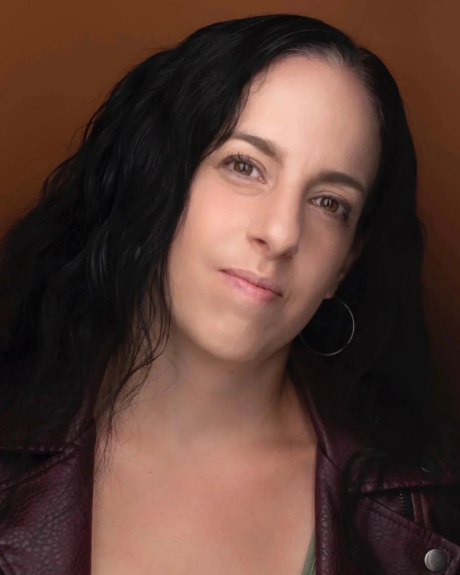 A woman with long dark hair, light skin, and hoop earrings, looking slightly to her right, with a soft smile, against a brown background.