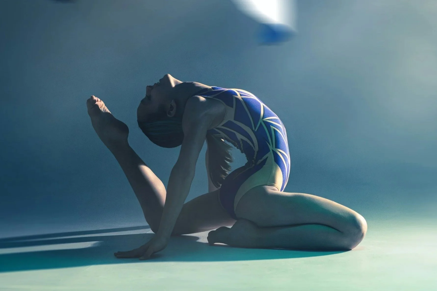 Gymnast in a blue and yellow leotard performing a floor routine pose on a light-colored mat, with a gradient blue background.