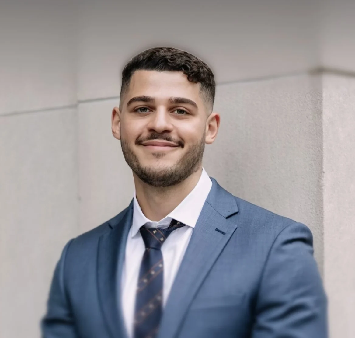 Portrait of a young man in a blue suit with a white shirt and plaid tie, smiling against a neutral background.