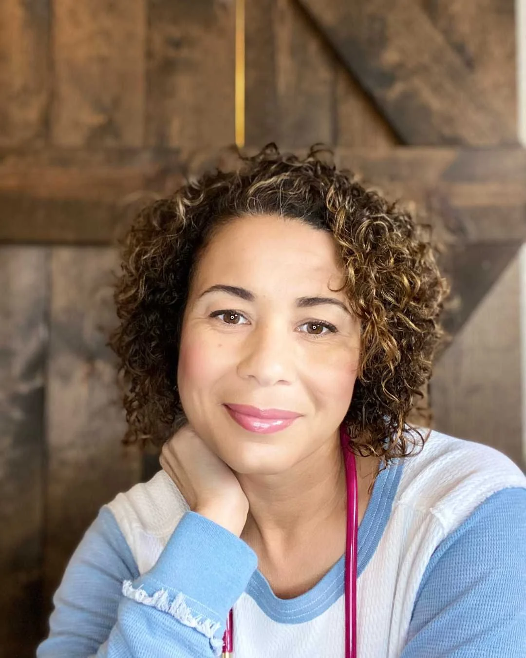A woman with curly brown hair smiling at the camera, resting her head on her hand, wearing a blue and white top with a pink stethoscope around her neck.