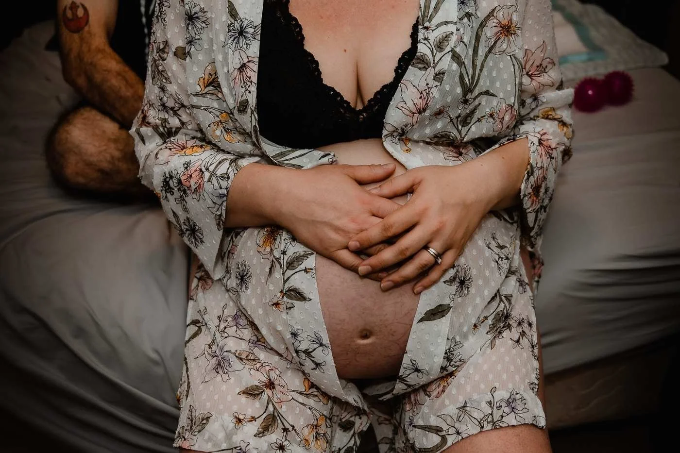 A woman in floral robes sits on a bed with her hands resting on her exposed belly, wearing a black lace top underneath; her partner, partially visible, is sitting behind her with legs crossed.
