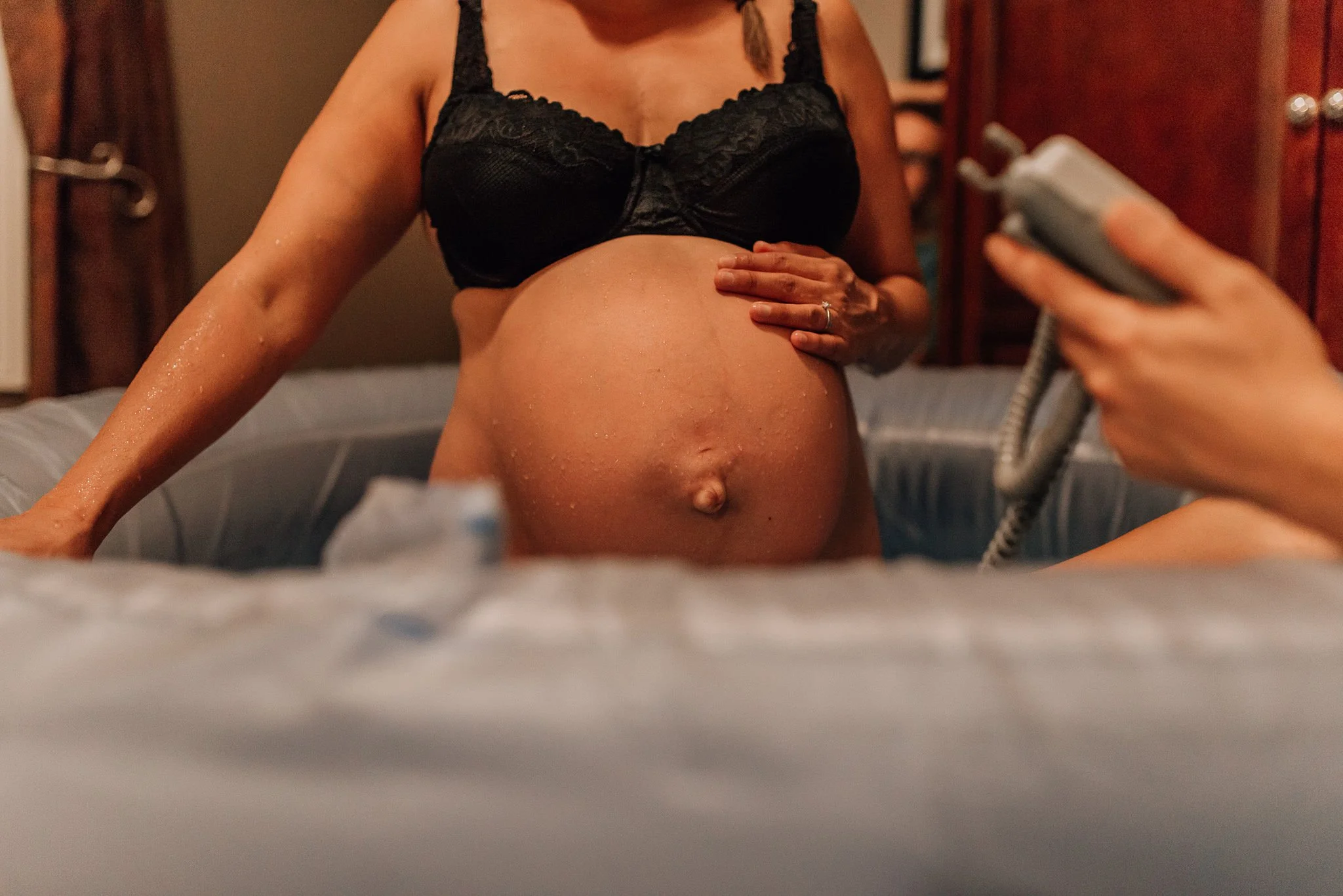 A pregnant woman in black lingerie sitting on a bed, holding her belly. Another person is partially visible, holding a landline phone to her ear. The scene appears to be in a bedroom with wooden furniture.