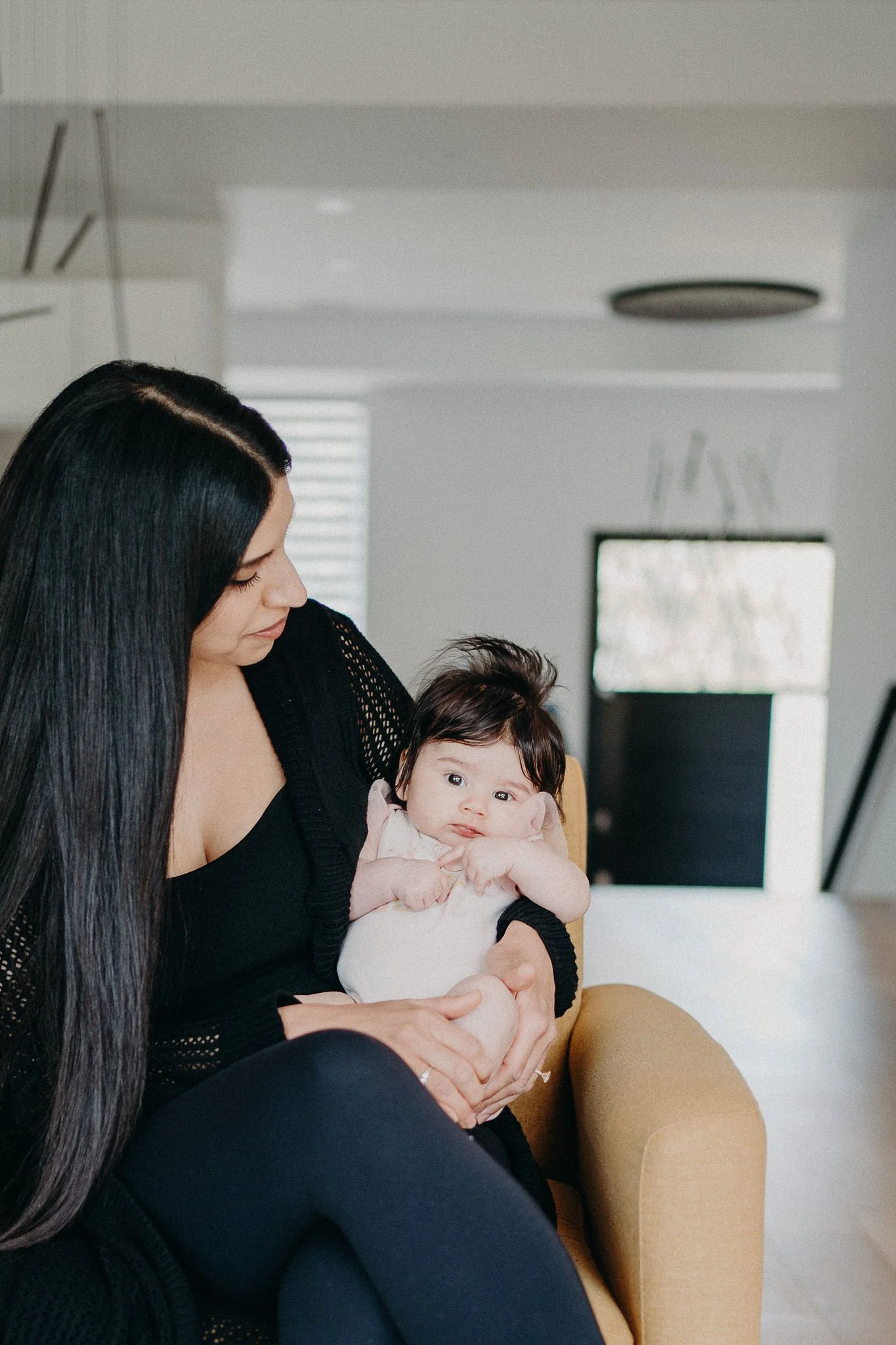 A woman with long dark hair holding a baby girl on a yellow chair in a modern living room.