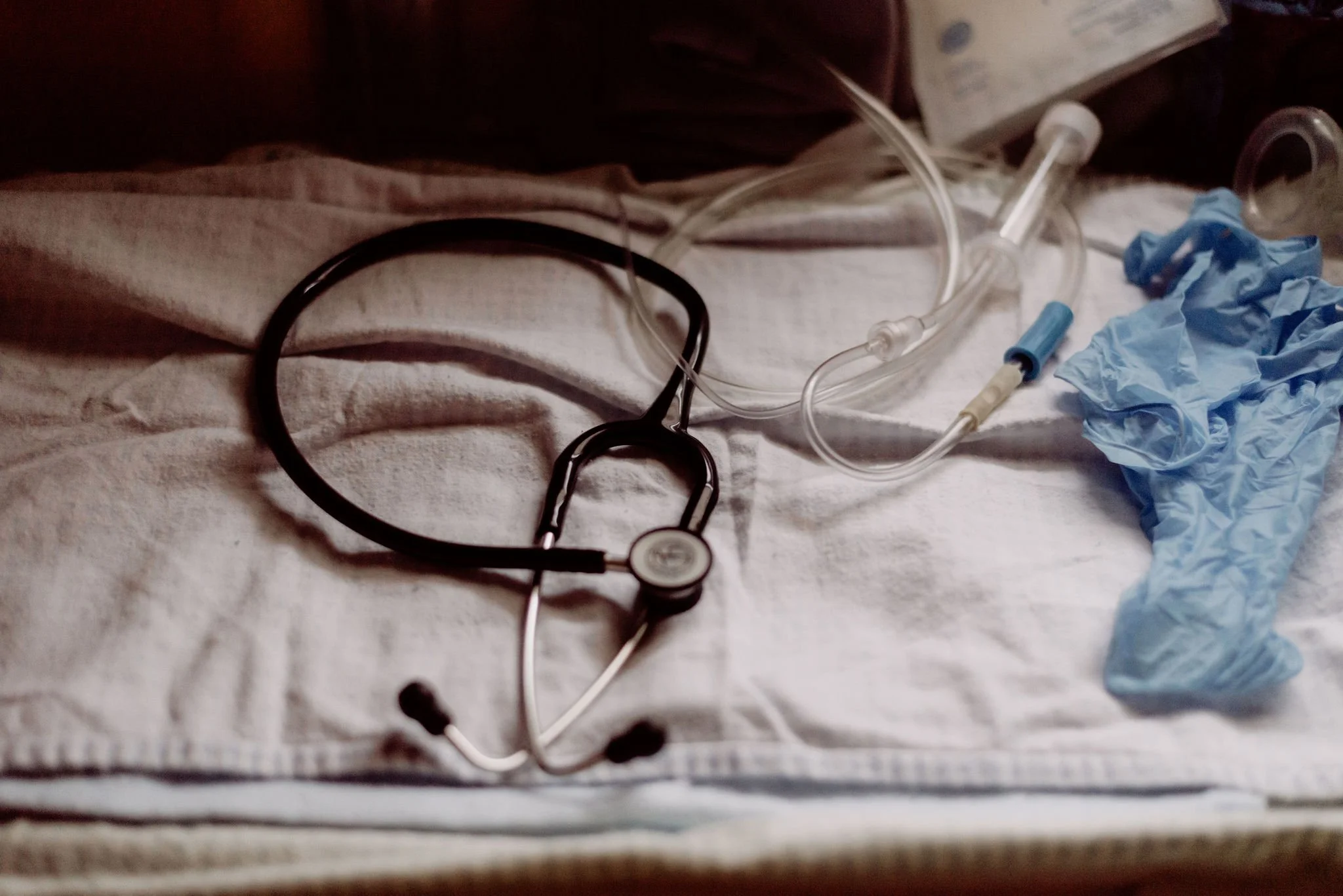 Medical scene featuring a stethoscope, nasal cannula, blue medical gloves, and a syringe on a hospital bed or medical tray.