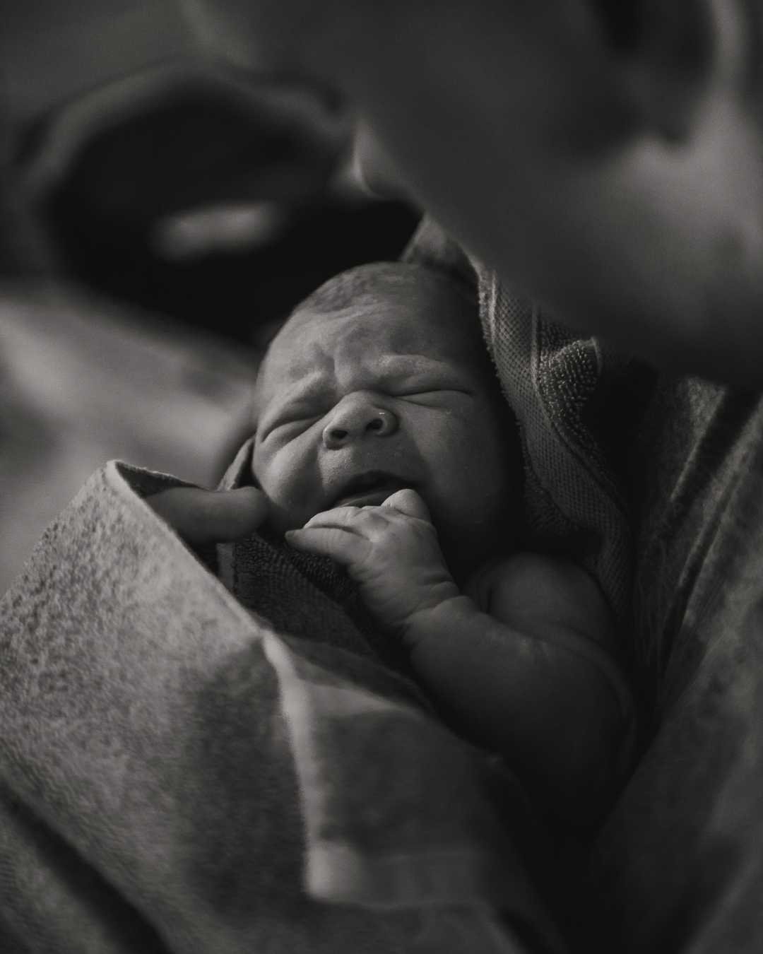 Black and white photo of a newborn baby sleeping peacefully, wrapped in a blanket and being held closely.