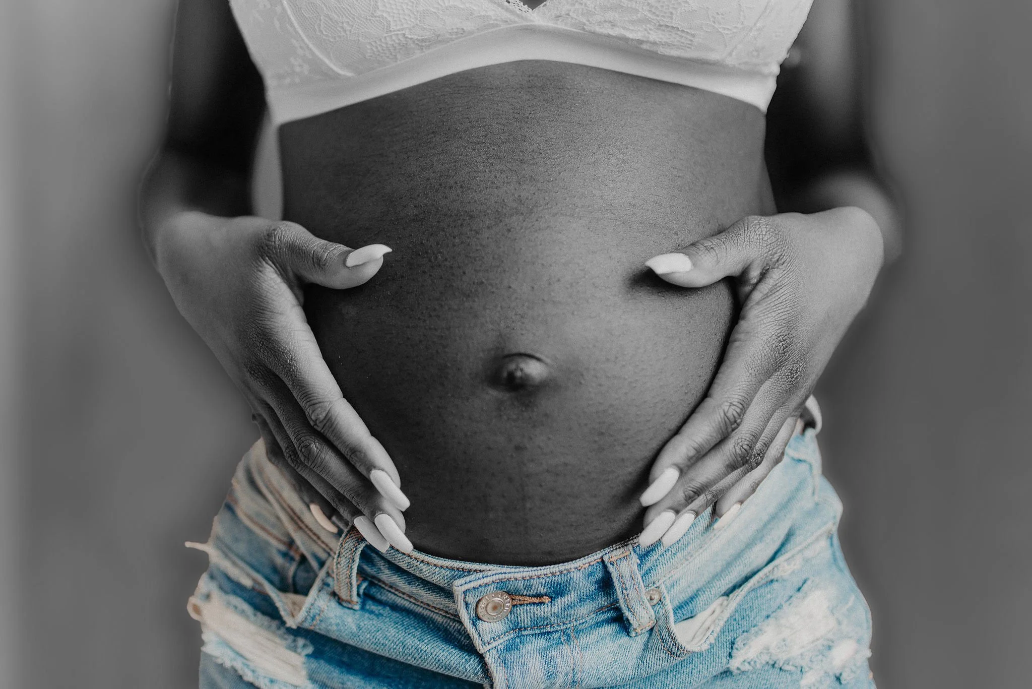 Close-up of a pregnant woman with dark skin, holding her belly with both hands, wearing a white lace top and ripped denim shorts, black and white photo.