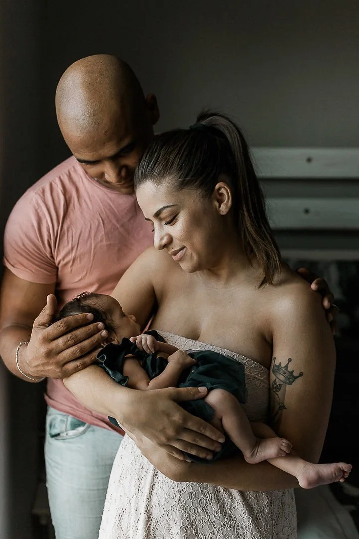 A woman holding her newborn baby, with a man standing close beside them, touching the baby's head, in a tender family moment.