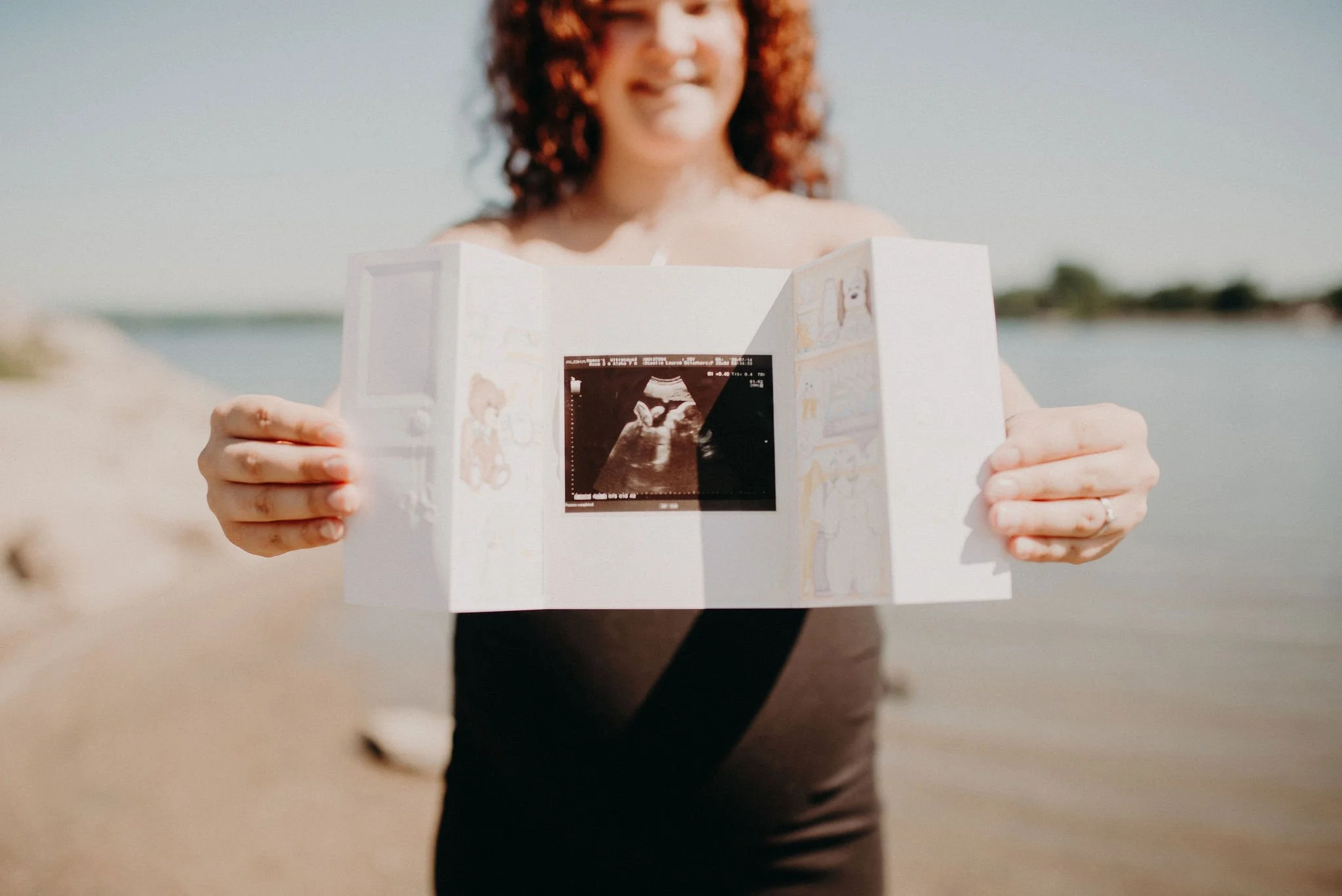 A woman holding a sonogram picture at the beach with water and a distant shoreline in the background.