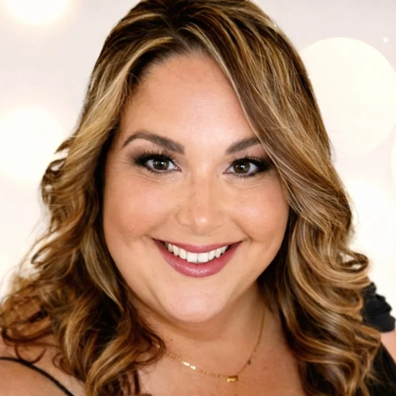 Close-up of a woman with wavy brown hair, smiling, wearing makeup and a black top with a gold necklace, in front of a blurred background.