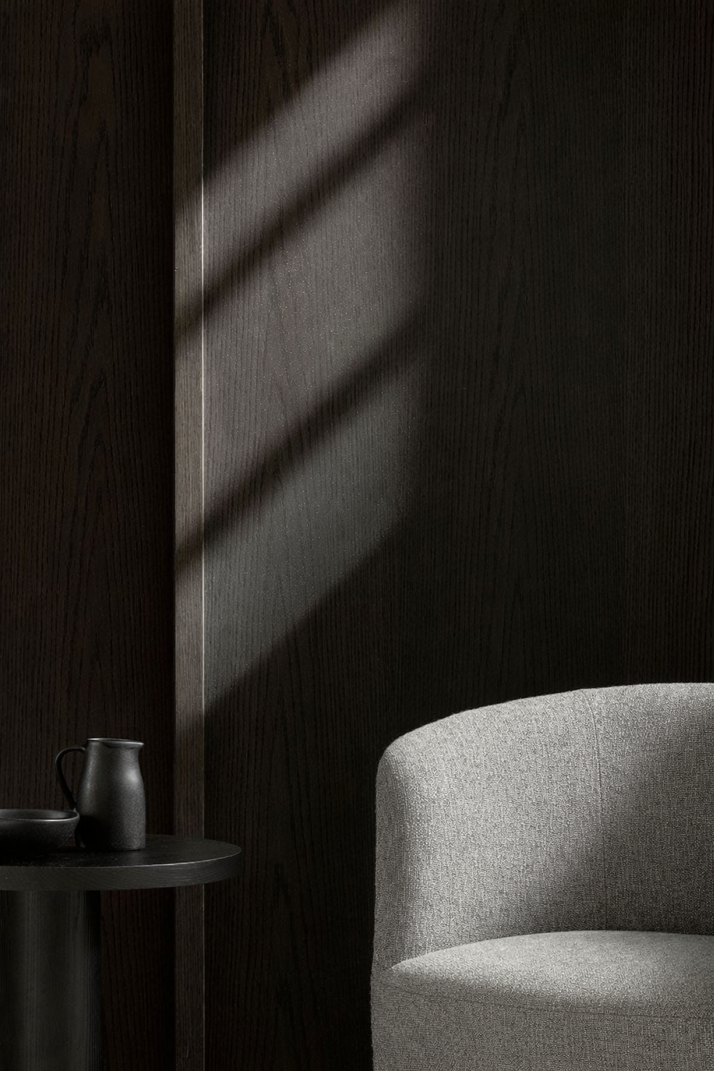 A cozy interior corner with a light gray upholstered chair, a small round black side table, and a dark wooden wall with sunlight casting diagonal shadows.