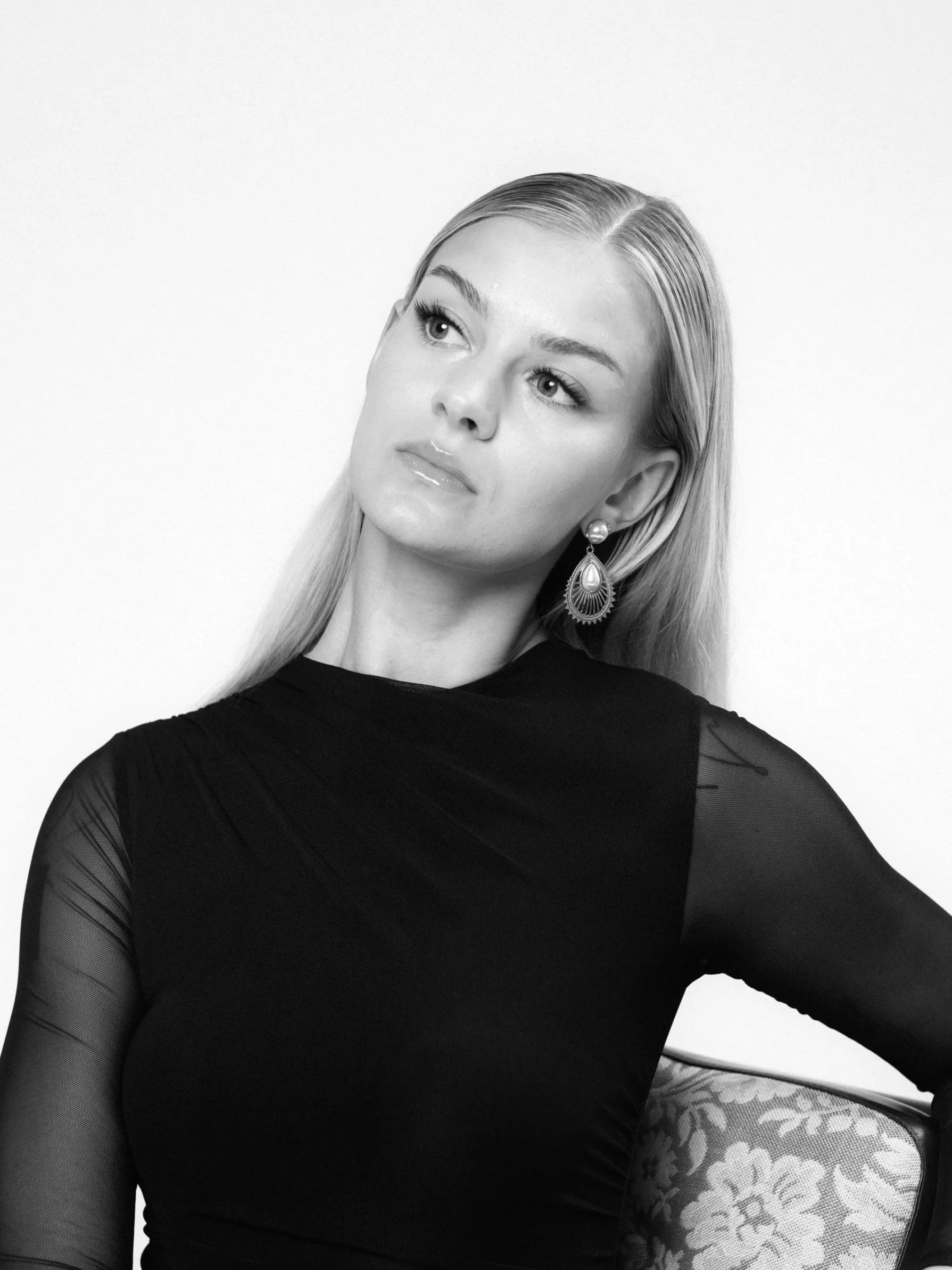 Black and white portrait of a woman with long blonde hair, wearing a black dress with sheer sleeves and ornate earrings, sitting on a patterned chair against a plain background.
