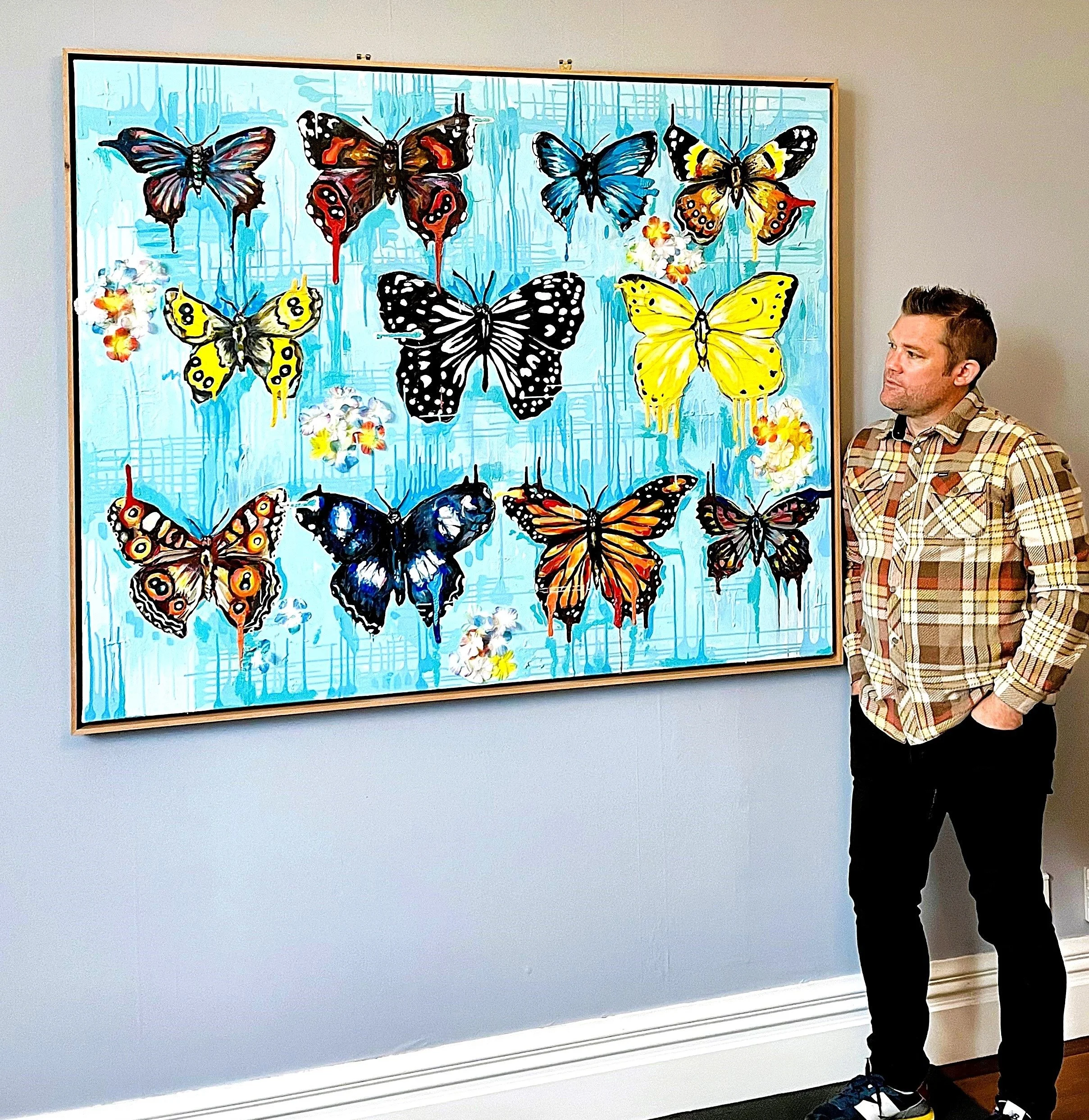 Simon Olsen in a plaid shirt stands with his hands in his pockets, looking at his colourful butterfly-themed painting hanging on the wall in a studio.